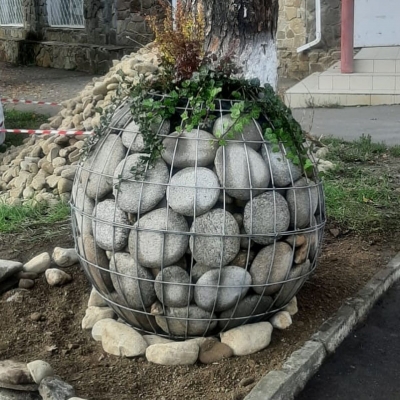 Шарообразная клумба из сварного габиона, наполненная крупными округлыми камнями и зелеными растениями.